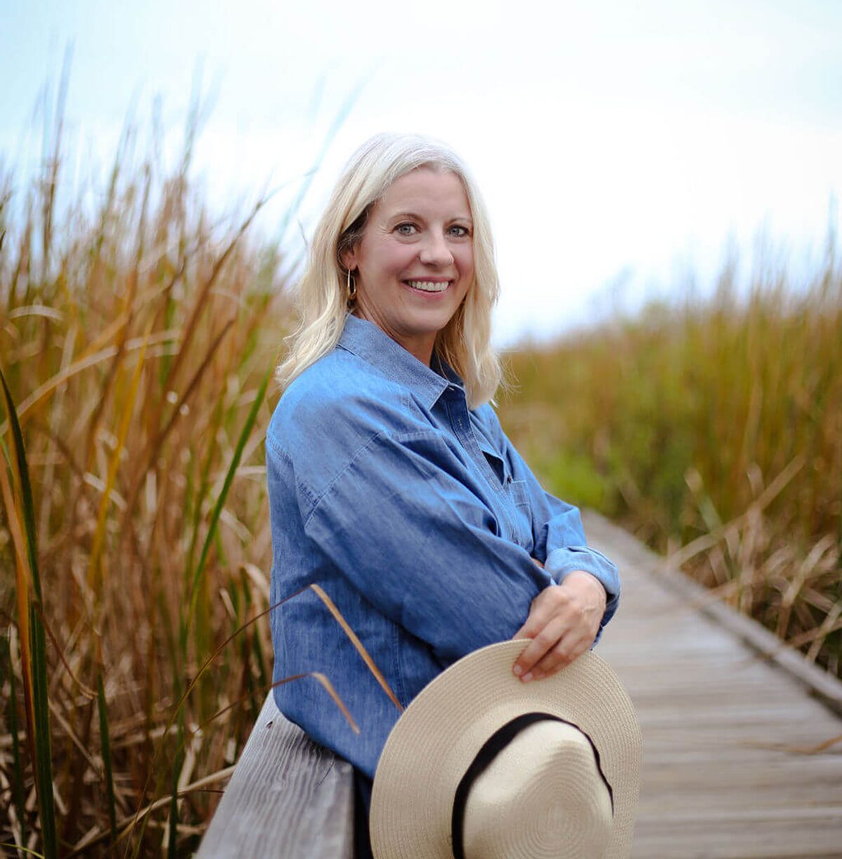 Jolene Park leaning on a beach boardwalk railing, holding a sun hat